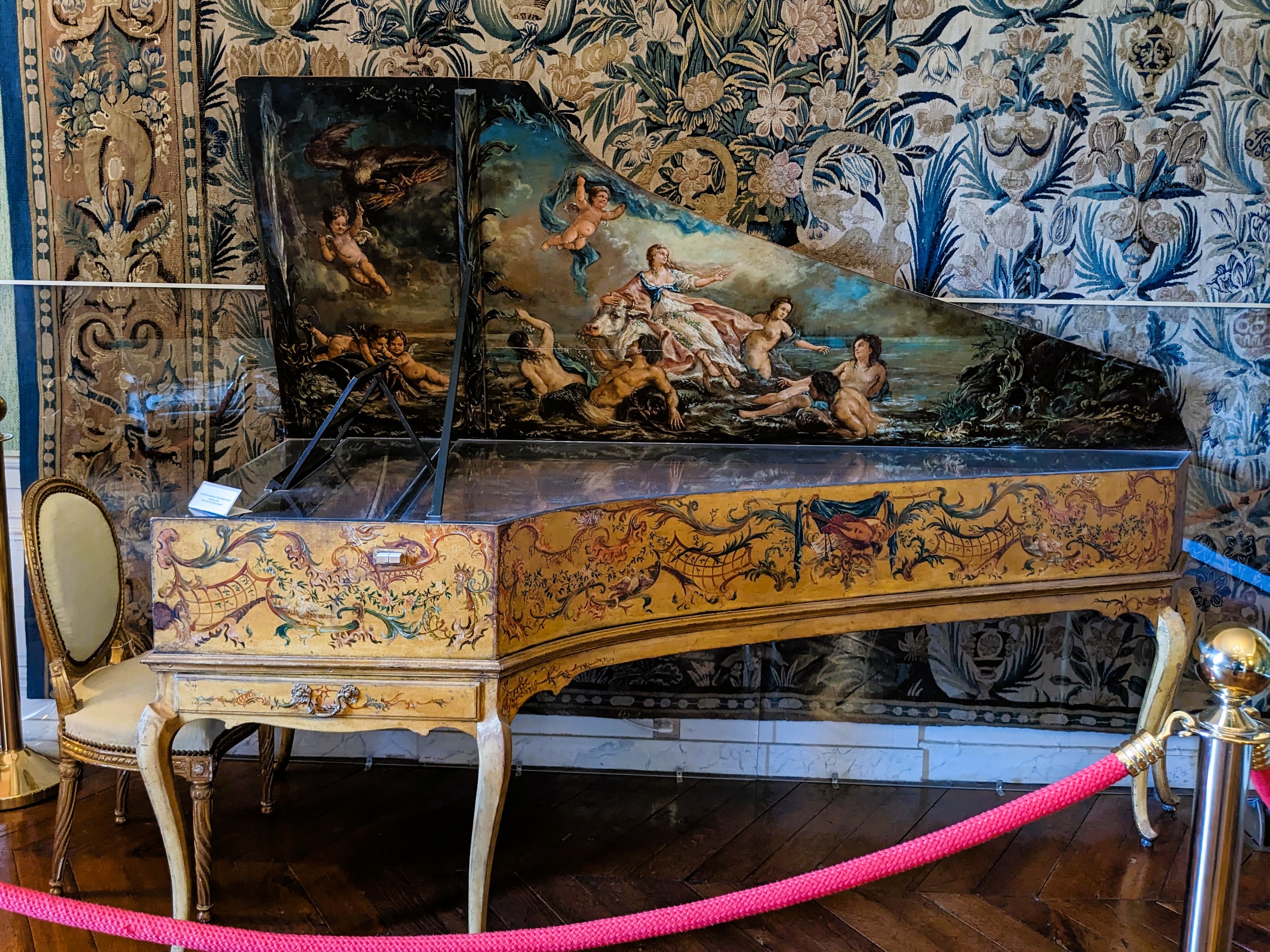Le clavecin d'Albert Delin daté de 1768 : caisse à décor peint de rinceaux, couvercle orné d'une scène mythologique, devant une tapisserie de verdure.